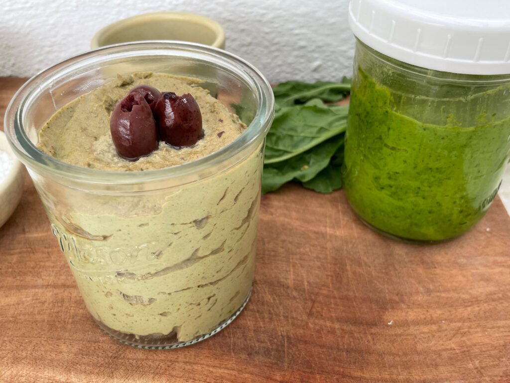 arugula hummus in a jar topped with olives in in front of a jar of arugula pesto on a cutting board with some arugula leaves behind them.