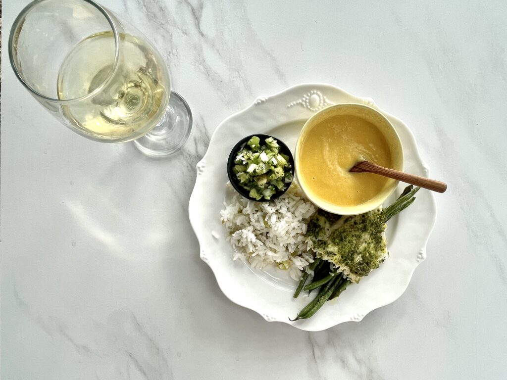 A light white table wine accompanies  dinner made up of arugula pesto crusted sea bass on a bed of green beans complemented by tomatillo rice, tomatillo salsa, and vegan creamy calabaza soup.
