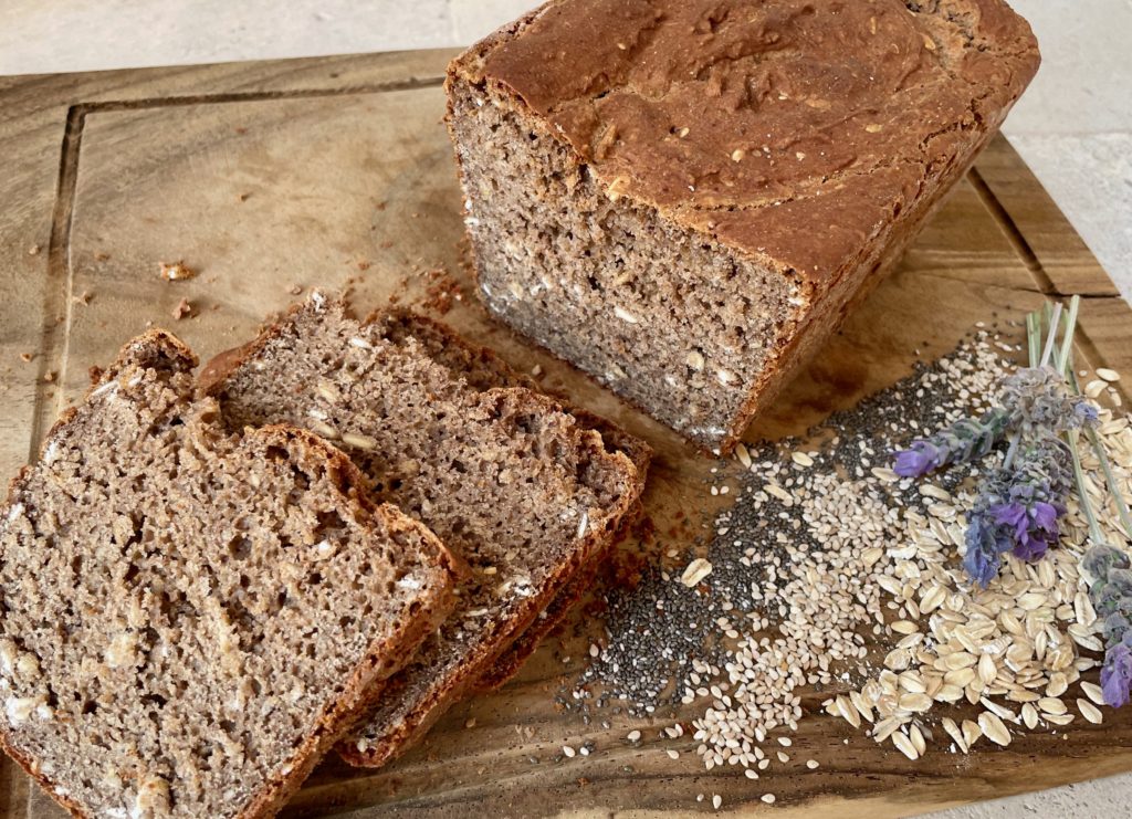 A crusty loaf of square bread rests on a wooden cutting board of a large size. The bread is dark brown, square, and crusty with many seeds and oats visible. Three slices are cut from the bread and stacked at an angle to the bread. A sweet arc of 3 inches is a rainbow of seeds, chia, oatmeal, sunflower seeds, and 2 lavender buds.A top view.