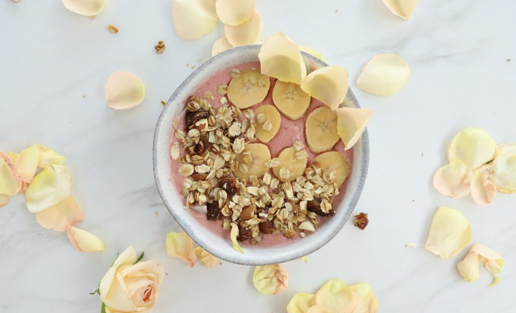 a blue pottery smoothie bowl with a littering of rose petals around the smoothie bowl covered with granola and bananas
