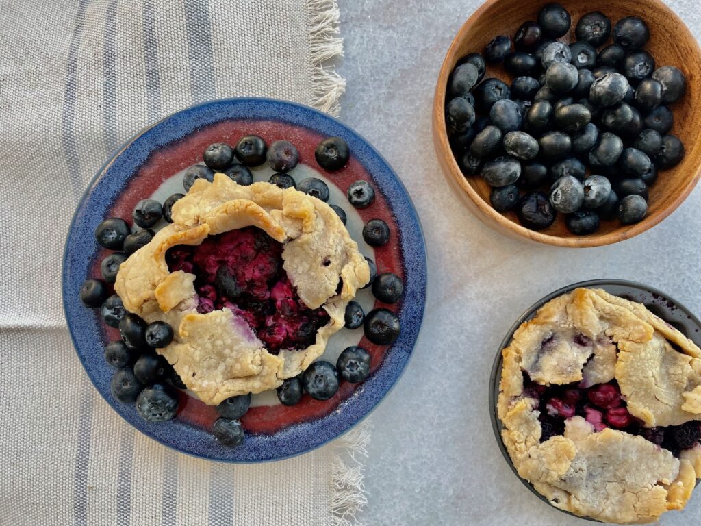 2 blueberry tarts with a wood bowl of blueberries is above a handwoven tea towel of linen and blue stripes