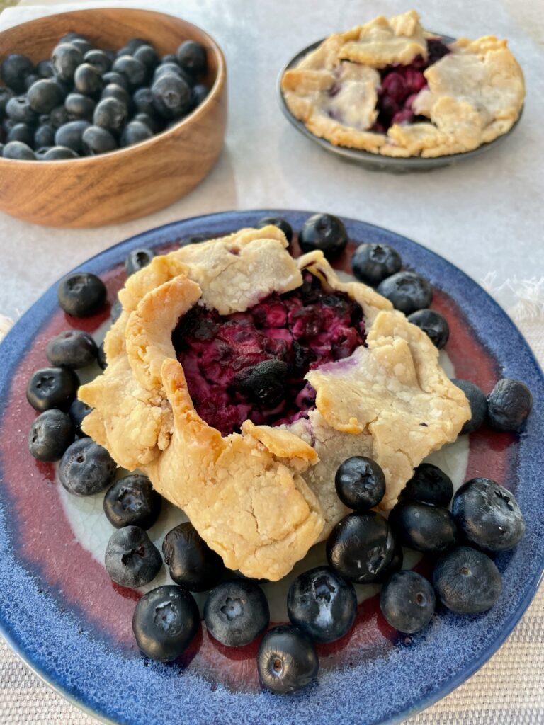 A close up of a round, free form blueberry tart or gallette is shown surrounded by fresh blueberries. 