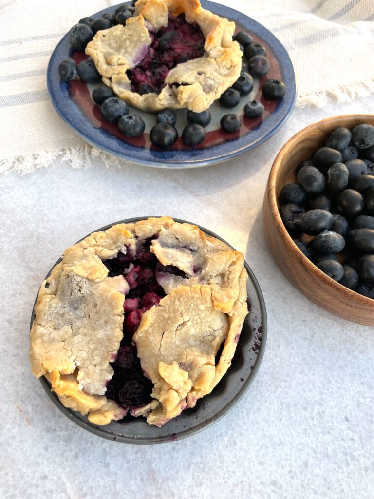 2 cooked blueberry tarts are shown next to a bowl of blueberries on a white marble background
