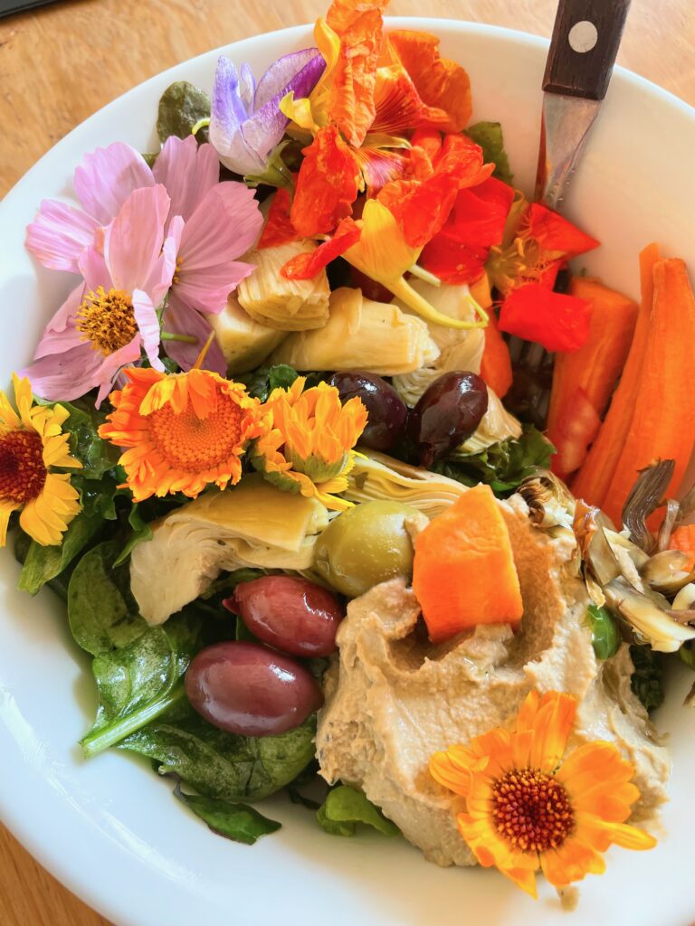 Edible flowers, hummus, artichokes, carrots, olives are above a bed of spinach and arugula