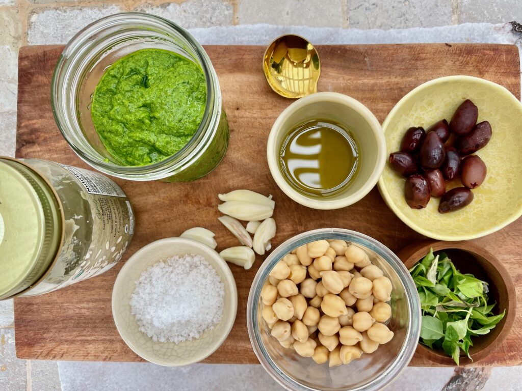 the ingredients for arugula pesto are shown...chickpeas, sunflower seed butter, olives, roasted garlic, fennel, salt, olive oil, olives