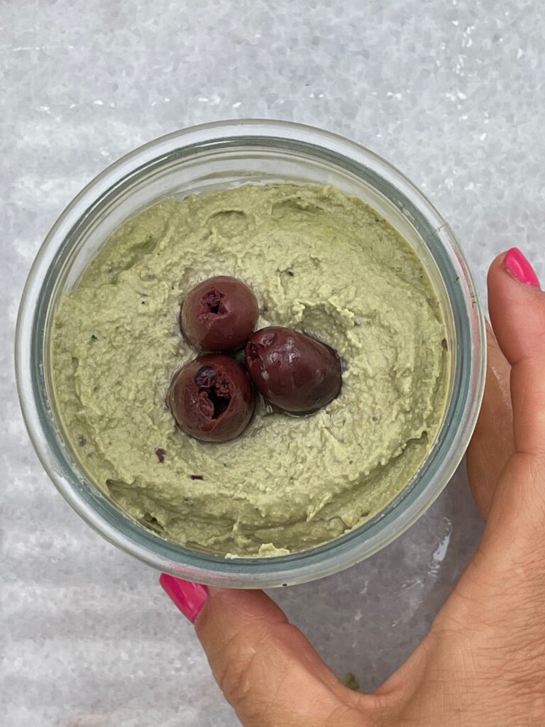 a hand with bright pink nails holds the hummus that is a glass jar and topped with a floret of black kalamata olives