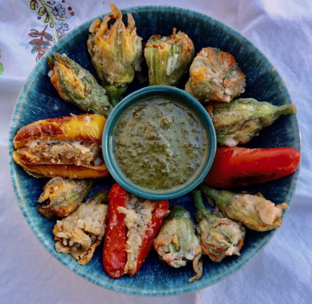 a blue pottery bowl has a basil pesto in the center and is full of stuffed squash blossoms, with 2 red peppers suffed at 3 and 6, and a yellow stuffed pepper at 9 o clock.