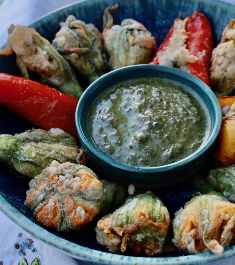 a close up cut off view of a blue pottery bowl has a basil pesto in the center and is full of stuffed squash blossoms, with 2 red peppers suffed at 3 and 6, and a yellow stuffed pepper at 9 o clock.