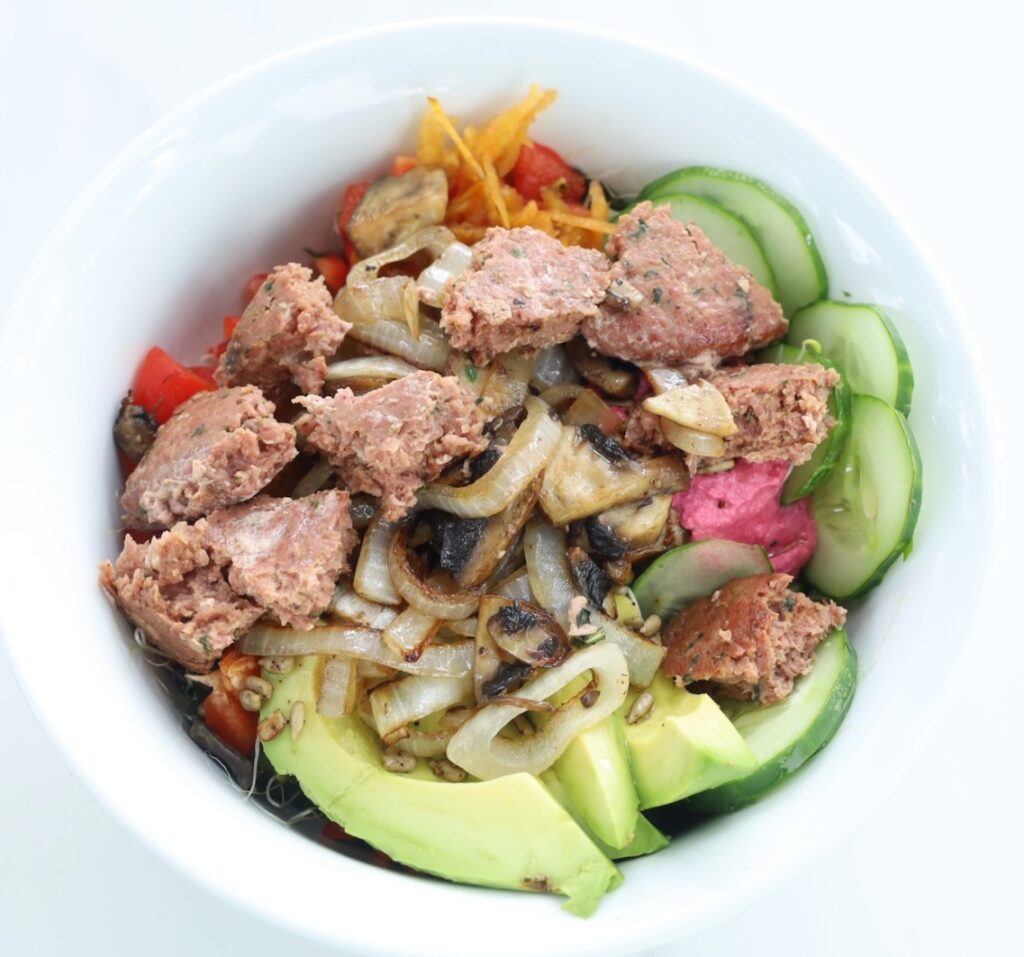 A circular arrangement of food showcases beautiful colors avocado, glimpses of beet hummus, red peppper, cucumber and yellow carrot. On top is a oily mound of caramelized onion and chunks of fried ground lamb meat. 