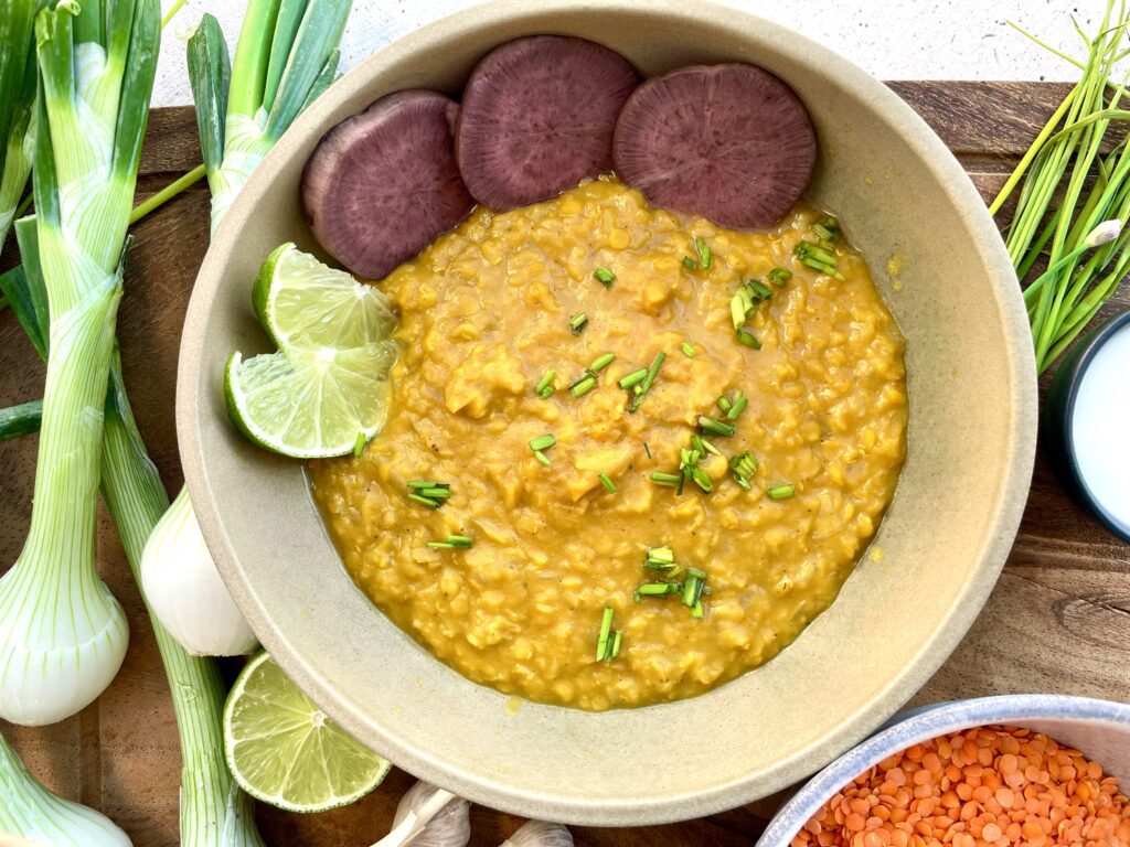 red lentil dahl is bright yellow colored and is in a bowl with three lentils slices and 2 lime wedges, surround with spring onions