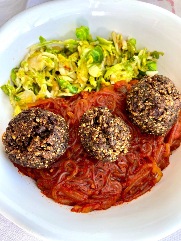 Three vegan meatballs are over a spagetti made with cellophane noodles and a side of sliced fried Brussel sprouts