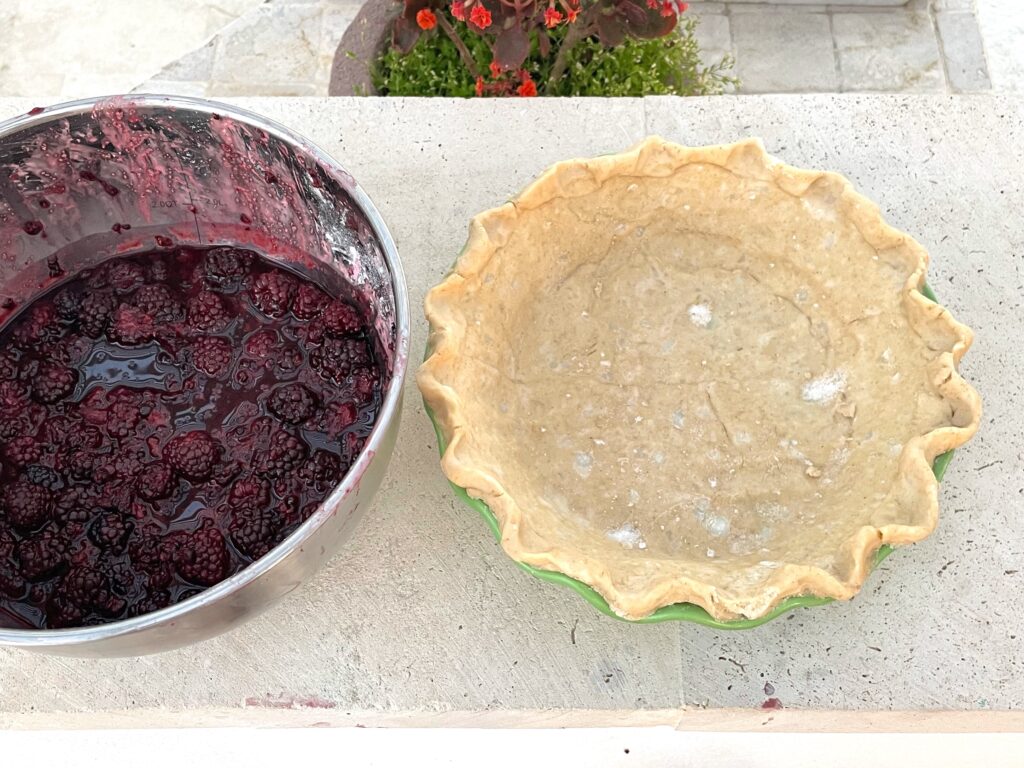 a large empty raw pie crust is shown next to a bowl of blackberry filling. The crust is in a green crockery dish and on a white stone background.