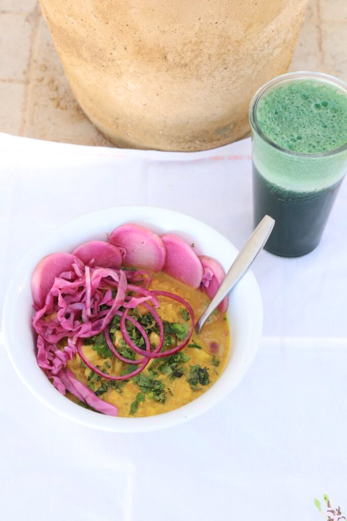 A bowl of cauliflower curry sits on a white background covered with pickeled daikon and cabbage...with a glass of spirulina pinapple juice in front of the base of a terra cotta pot.