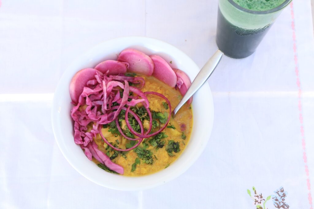 A bowl of cauliflower curry sits on a white background covered with pickeled daikon and cabbage...with a glass of spirulina pinapple juice in on a white background