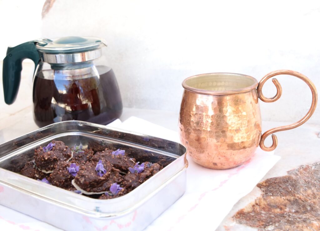 a stainless steel box of chocolates is featured from the side angle with the background containing a glass tea pot of dark red liquid, hibiscus tea next to a bright copper mug. 