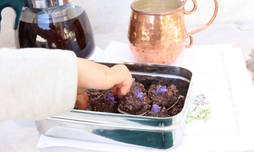 a stainless steel rectangular box hold around 21 mounds of chocolate truffles each topped with a purple lavender flower. A child's hand reaches into the box to select a chocolate.