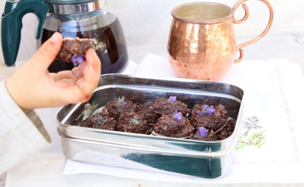 a stainless steel rectangular box hold around 21 mounds of chocolate truffles each topped with a purple lavender flower. A child's hand holds one truffle face up.