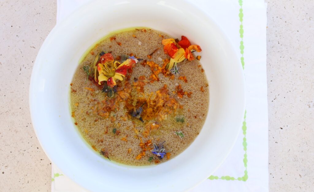 A large sloping white bowl is filled with amaranth porridge and topped with fresh edible flowers, a grate of turmeric and coconut oil that is melted.