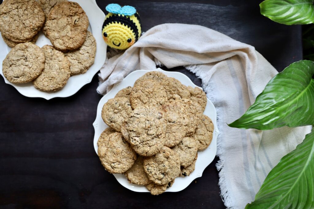 two white plates contain yummy cookies that are 3 inches in diameter. They are resting on a black stained table and a textile cotton cloth of natural fiber is draped around the plate to the position of 3 o clock. The other plate is at 12 o clocK. A stuffed bumble bee is at 1 o clock. A few plant leaves have made their way into the picture