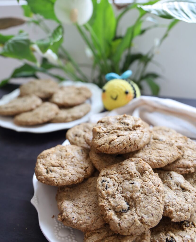 a plate of textured brown cookies are 3 inches across and on a white plate on a dark table with a second plate, a knit bumble bee and a lily of the valley plant in the background which are not in focus like the front bee.