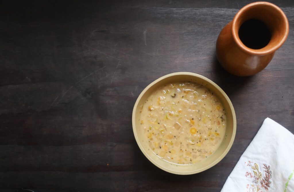 A light tan pottery bowl contains a soup that is similar in color to the bowl and is flecked with small rounds of carrot and squares of cabbage. A white linen napkin embrowderied wiht bluebonnets is on the side and a tall clay drinking vessel is shown at the top right corner.