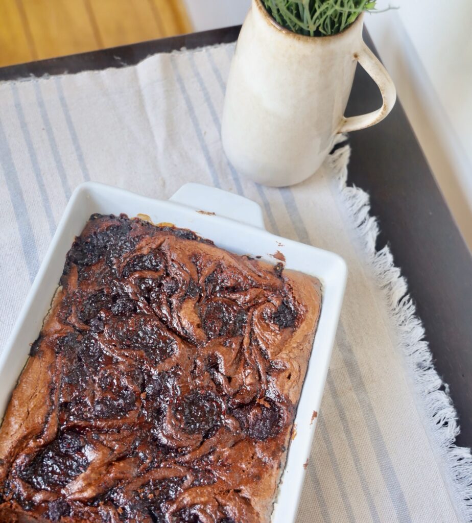 a tray of brownies is in a white mason dish with a background of a linen pale blue striped tapestry. A beige vase is on the corner of the black table that holds the brownies. 