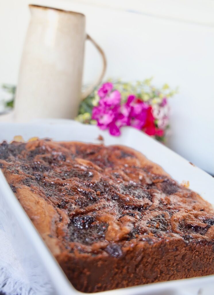 A side shot of the berry brownies in a white mason dish, with the edge cut so you can visualize the rich dense body of the brownies. A spattering of tiny fushia pink flowers are in the back corner bunched next to a beige pottery vase. 