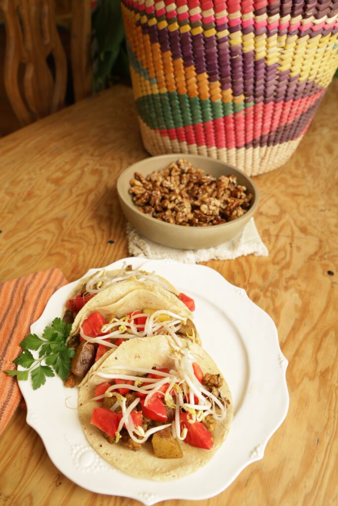 a plate of tacos with bright red tomatoes and sprouts are shown in fornt of a bowl of molasses walnuts in front of a large handmade colorful basket.