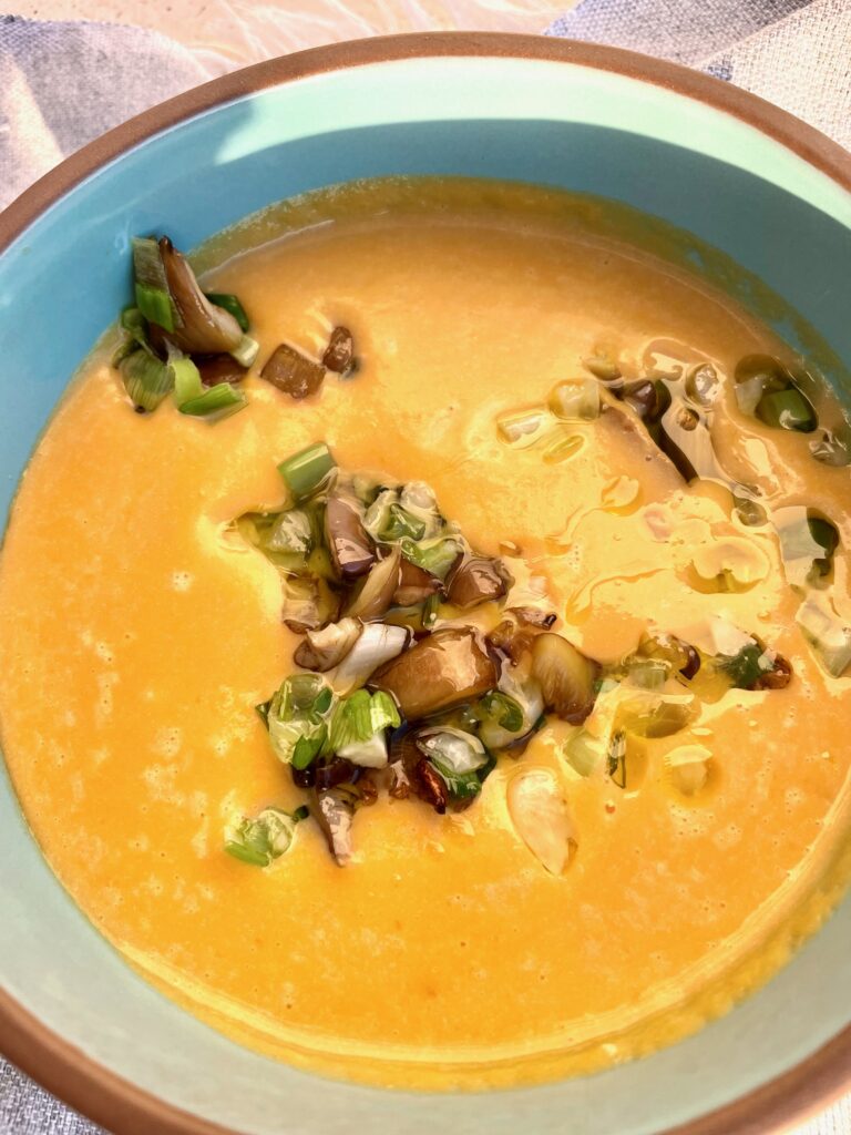 a blue brown rimmed pottery bowl contains a bright yellow soup that is topped with olive oil, fried mushrooms and green onions. 