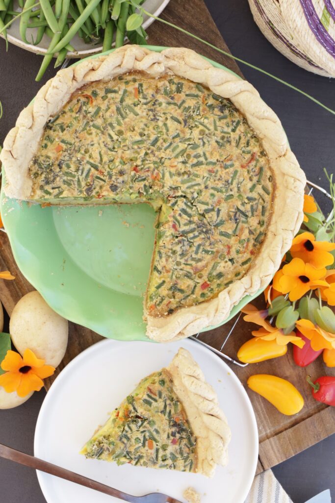 A vegetable quiche in a green pottery pie dish is surrounded by orange flours, bright red peppers, and potatoes. A slice is cut out of the whole pie and put on the side.