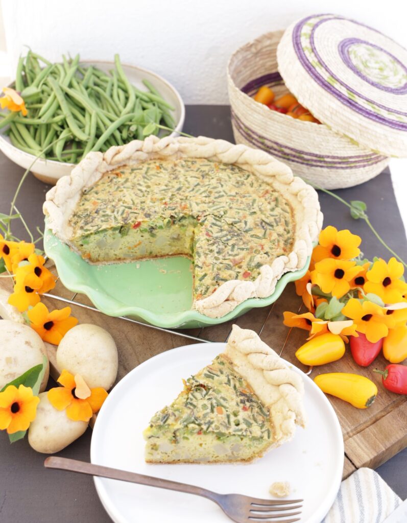 A vegetable quiche in a green pottery pie dish is surrounded by orange flours, bright red peppers, and potatoes. A slice is cut out of the whole pie and put on the side. A large plate of green beans and a hand woven basket is shown in the background.