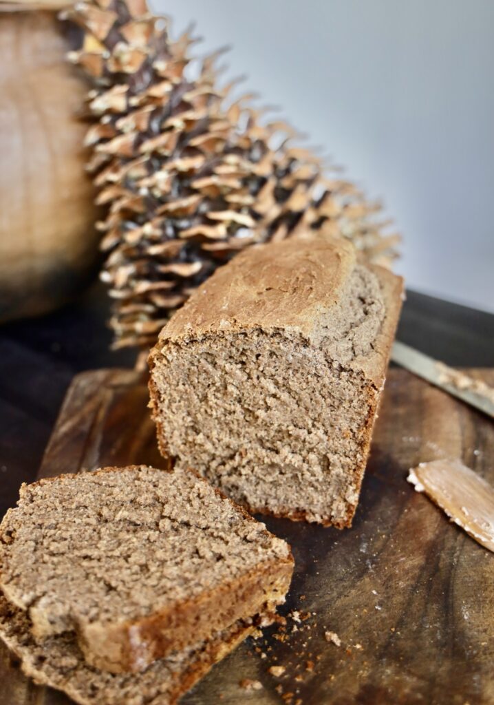 A loaf of brown bread is sliced on a cutting board in front of pine cones and a large clay pot. 