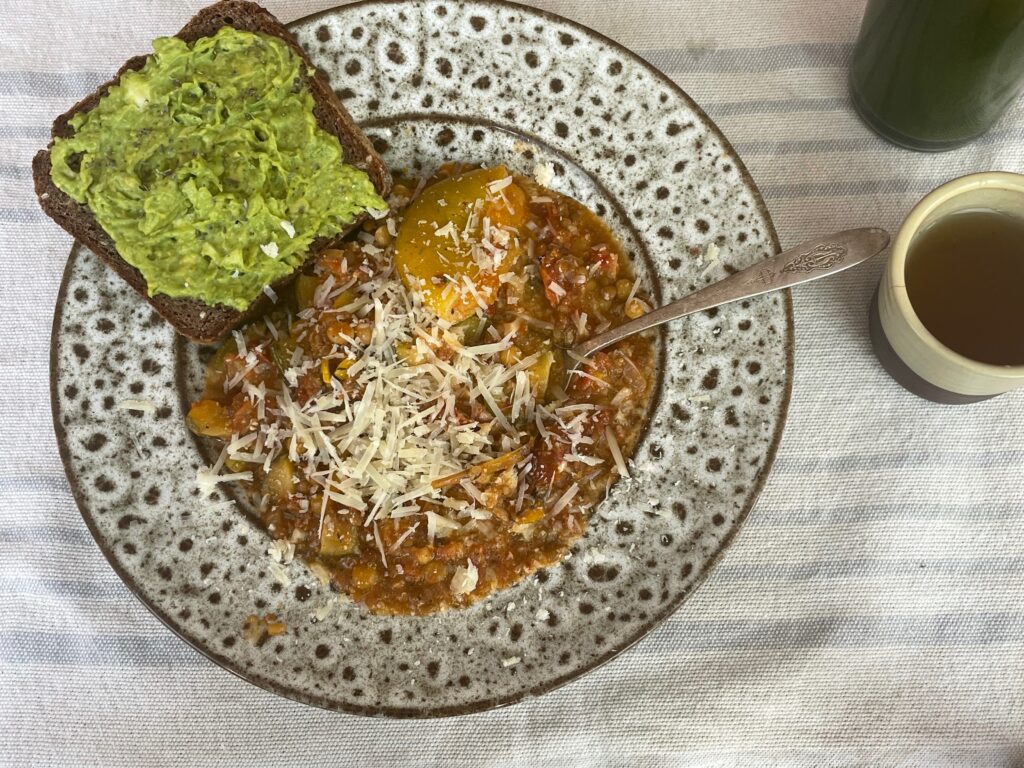 A bowl of the summer stew is in a large speckled egg dish and topped with parmesan cheese with a side of teff avocado toast. A side of green tea rests next to the silver spoon.