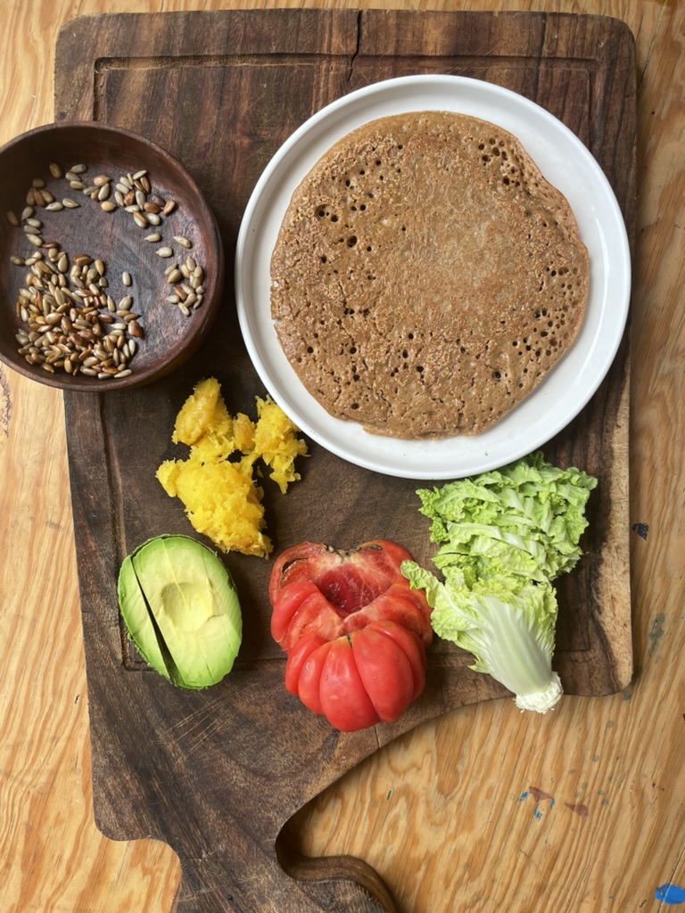 A cutting board contains ingredients for the wrap, the wrap, the avocado, the tomato, the squash, and a lettuce head, and a bowl of seeds.