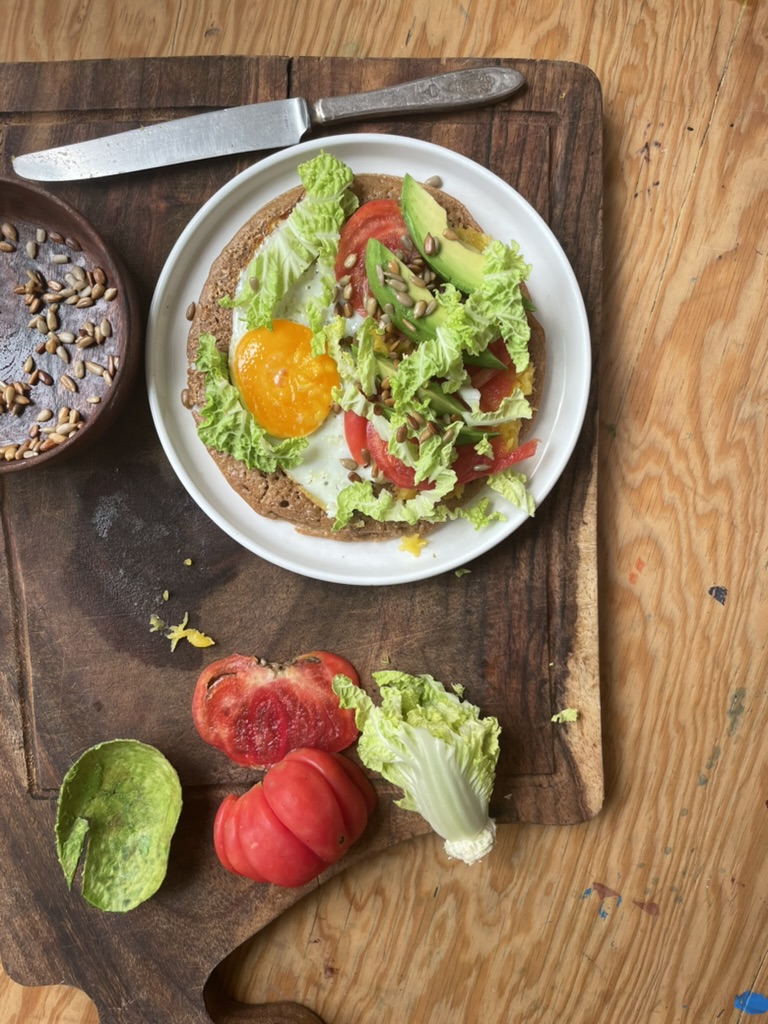 A cutting board contains ingredients for the wrap, the wrap, the avocado, the tomato, the squash, and a lettuce head, and a bowl of seeds.