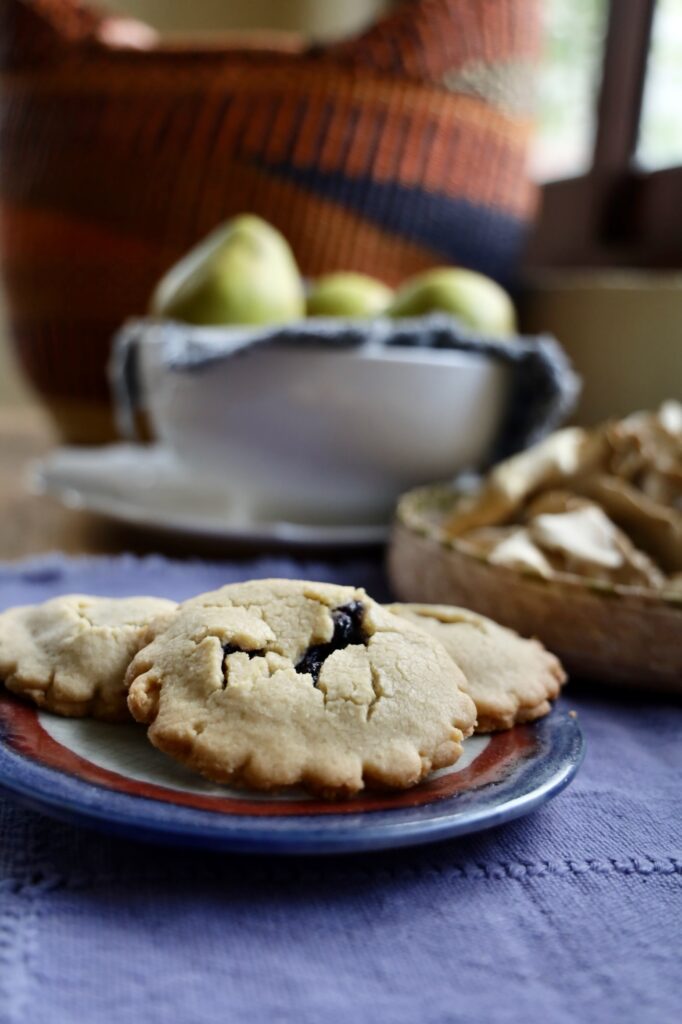 A colored pottery plate shows three jam filled cookies with a a purple textile background and a basket of dried mushrooms, a bowl of pears, and a rattan basket in the back ground on a wood table in front of a window.