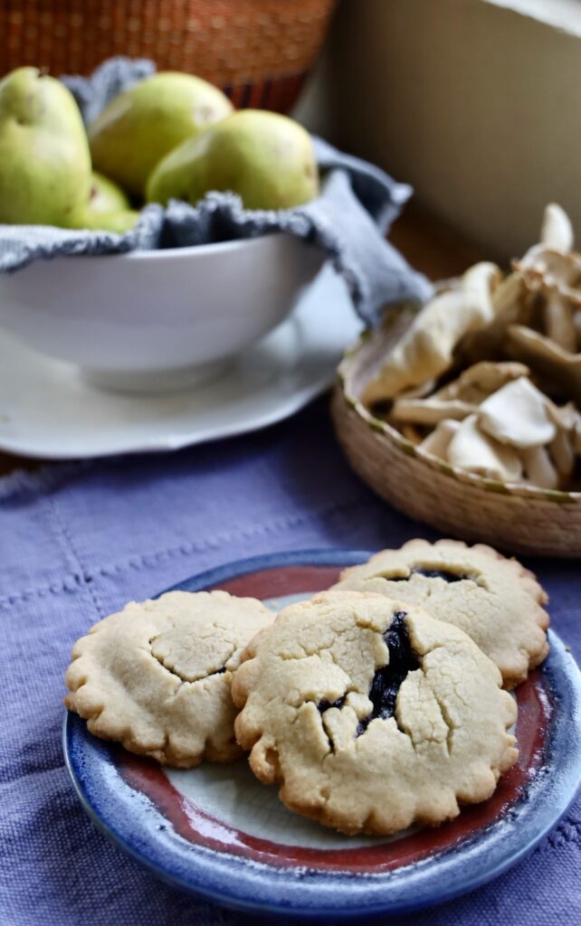 A colored pottery plate shows three jam filled cookies with a a purple textile background and a basket of dried mushrooms, a bowl of pears, and a rattan basket in the back ground on a wood table in front of a window.