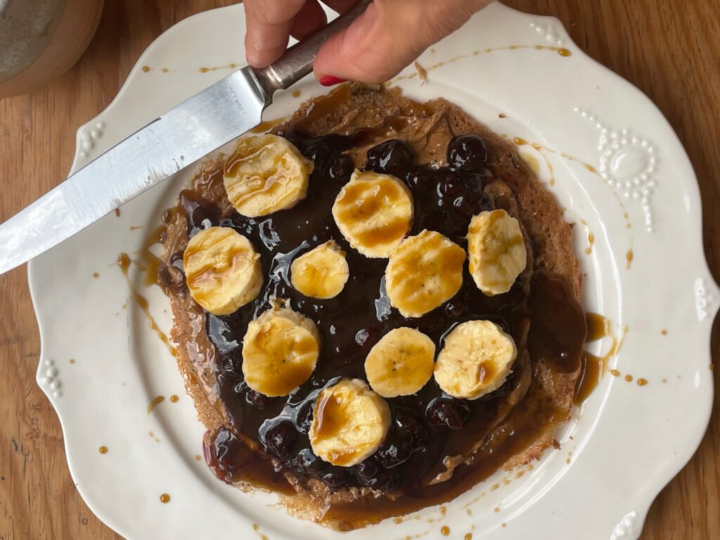 an almond teff wrap, covered in almond butter, blueberry compote, sliced bananas, and covered with blackstrap molasses. a hand holds a silver butter knife ready to cut a bite.