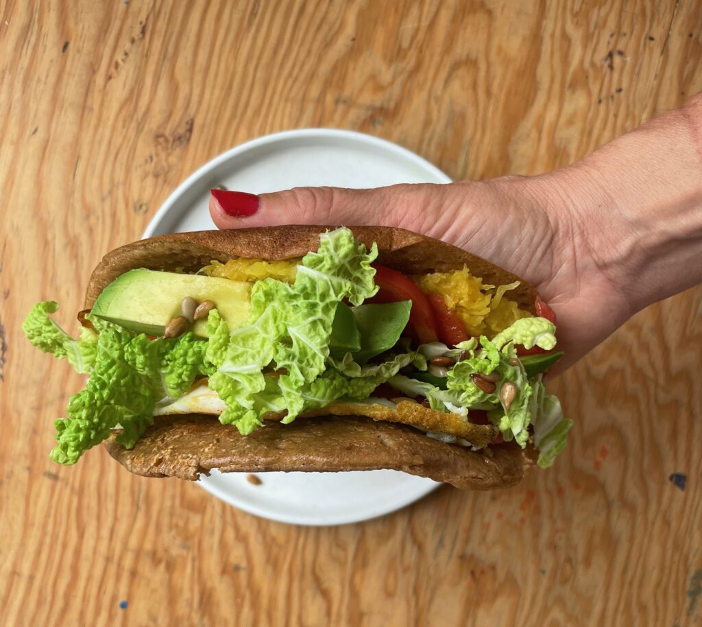 A polished hand holds a wrap filled with egg, avocado, lettuce, tomato, and sunflower seeds