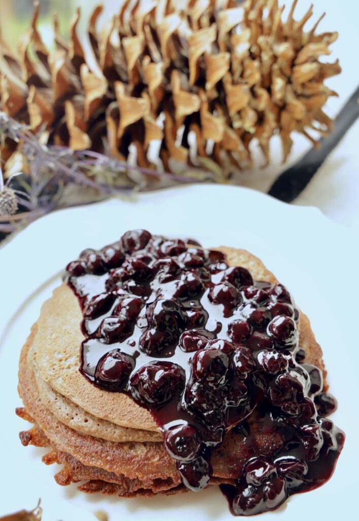 A stack of Blender Pancakes is covered with a large amount of Blueberry Jam Compote on a white plate. The angle is from the top of the pancakes that are on a white plate.