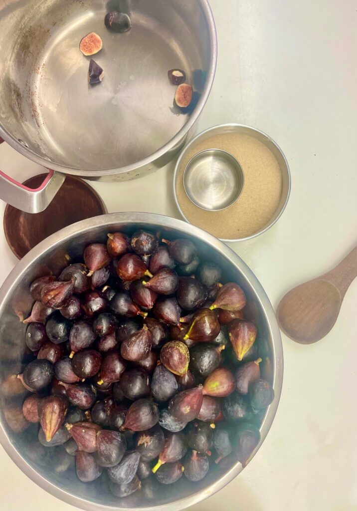 The ingredients are laid out: a large bowl of figs, the paot they will be sliced into, a knife, a sweetner, and a wooden spoon.