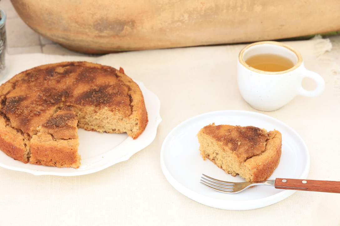 A slice of Maca cake is on a small lipped white plate with a fork. To the upper left of this slice is the whole cake with the slice missing. It is round and crusted with a dappling of brown sugar. To the upper right of the slice of cake is a cup of green tea in a thick bulbous white mug. A long, oblong, clay pottery pot is behind the place setting.