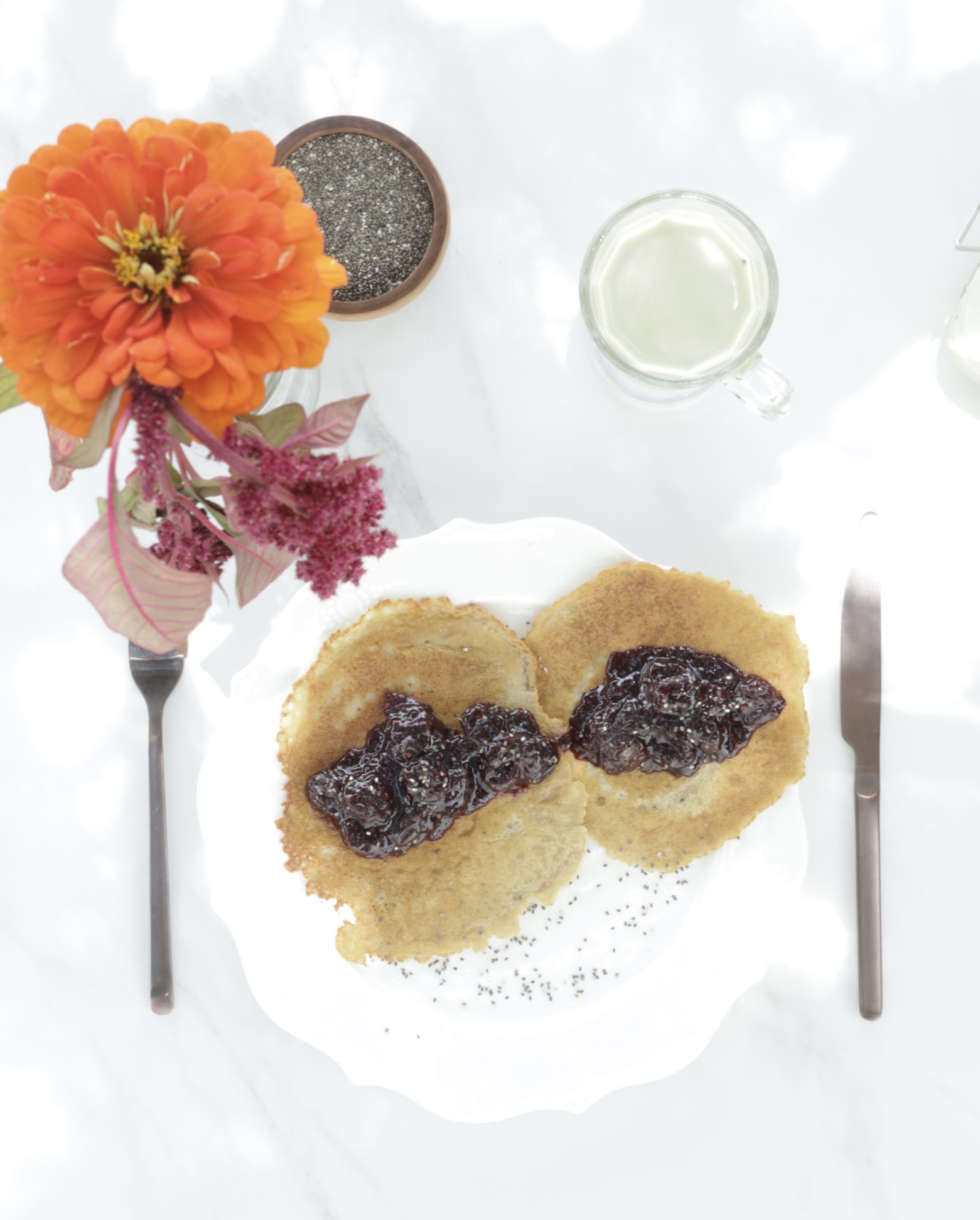 traditional crepes with a filling of cherry chia compote plated on a white plate in a place setting with zinnias and a delicate cup of matcha