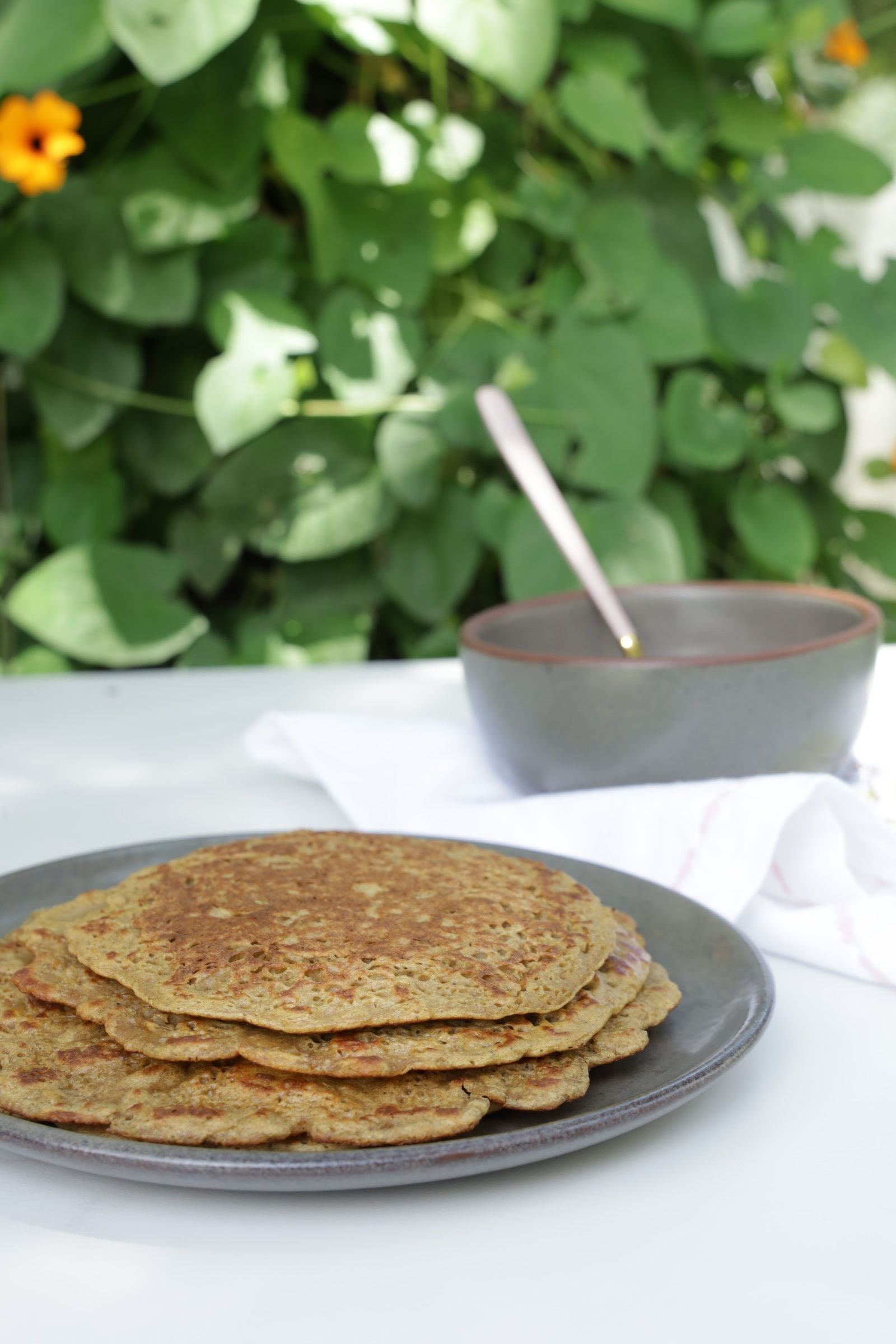 A stack of whole grain crepes in front of a grey pottery bowl and a pink spoon in front of a vine with orange flowers