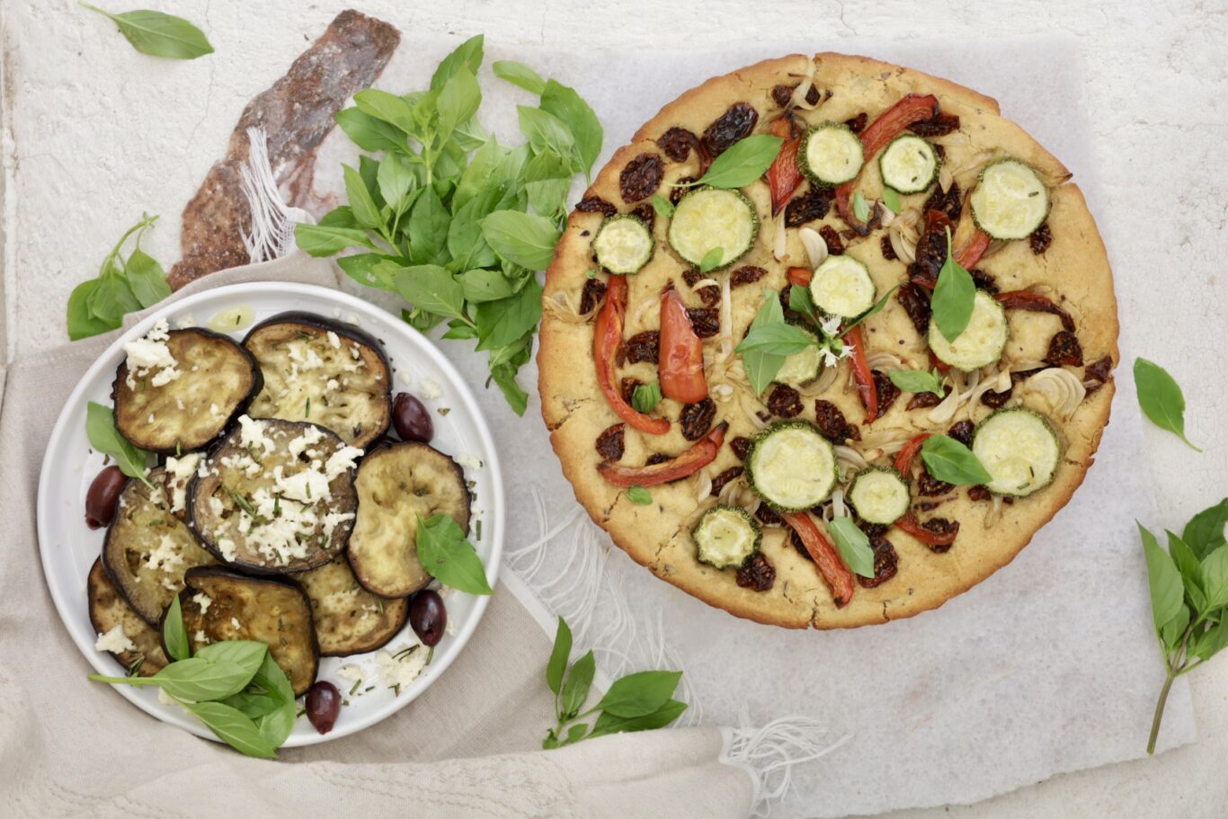 There are two plates. One has the gluten-free focaccia bread covered with zucchini, peppers, olives and fresh basil leaves. The other plate is white and is plump with a pyramid of roasted eggplant. fresh basil and a linen towel are interspersed with the plates.
