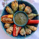 a blue pottery bowl has a basil pesto in the center and is full of stuffed squash blossoms, with 2 red peppers suffed at 3 and 6, and a yellow stuffed pepper at 9 o clock.