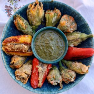 a blue pottery bowl has a basil pesto in the center and is full of stuffed squash blossoms, with 2 red peppers suffed at 3 and 6, and a yellow stuffed pepper at 9 o clock.