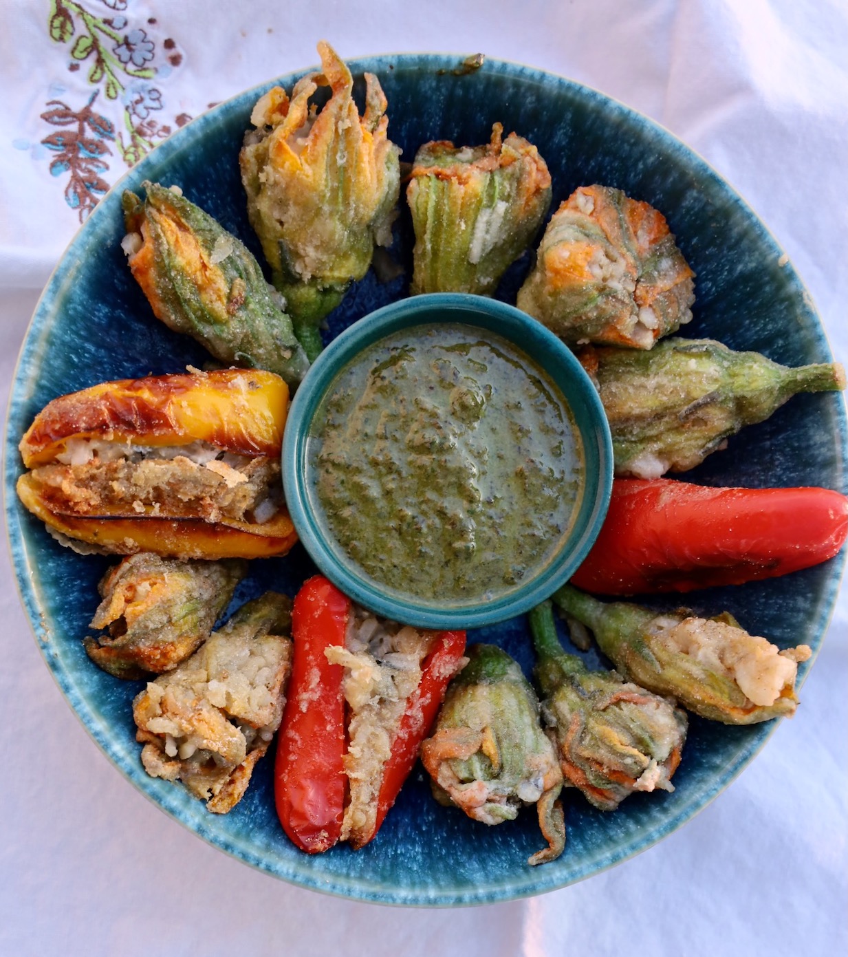 a blue pottery bowl has a basil pesto in the center and is full of stuffed squash blossoms, with 2 red peppers suffed at 3 and 6, and a yellow stuffed pepper at 9 o clock.