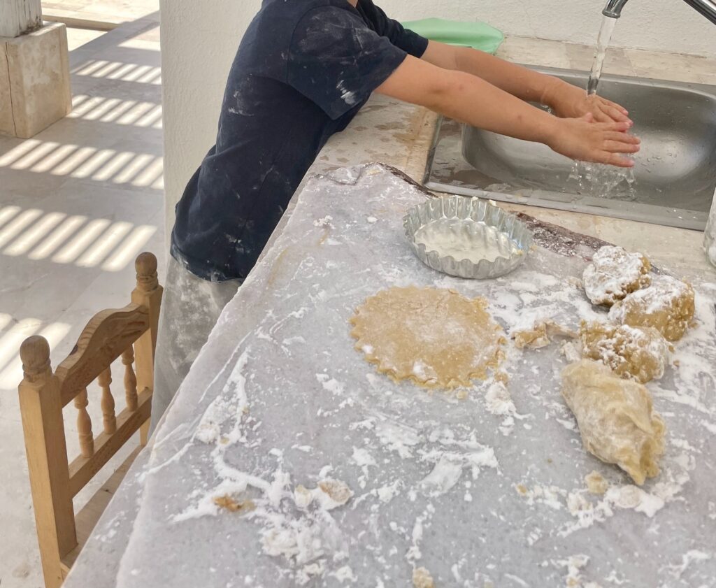 a marble slab is sprinkled with tapioca starch, dough, and cookie cutters. A little boy washes his hands in the background and you see his