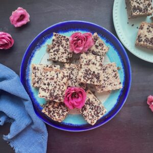 an angular arrangement of square bars sit atop a brilliant blue pottery plate. The cookie bars are mottled wiht cacao chips and two roses that are brilliant pink are nestled in the bars. two plates are whowen and the backround is black wood, a cotton neutral blue tea towel is the the left of one of the plants. the other plate is in the top right courner and you only see 1/3 of the plate.